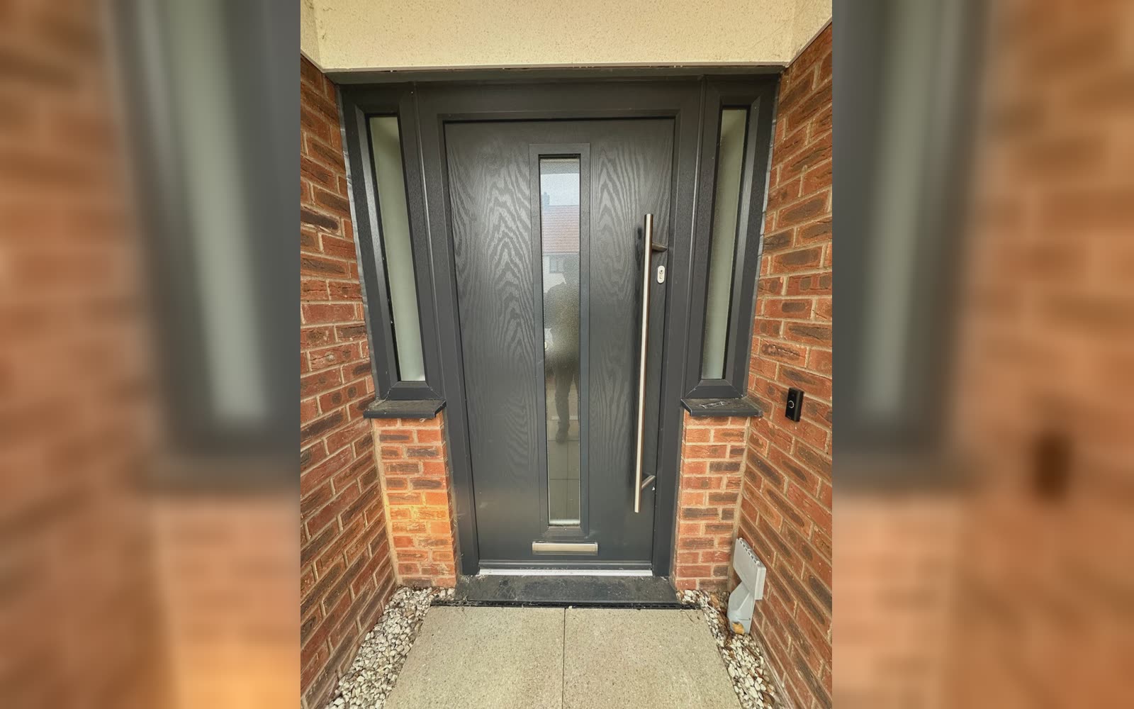 An anthracite grey composite front door with a long brushed steel handle and matching glazed side panels, fitted by URG in a red brick house